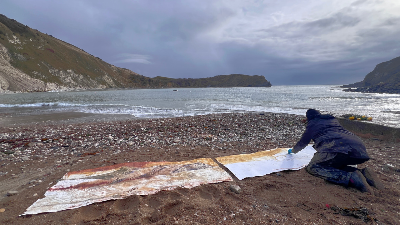 Frances kneeling at the tide line at Lulworth Cove, working on a long painting laid on the beach
